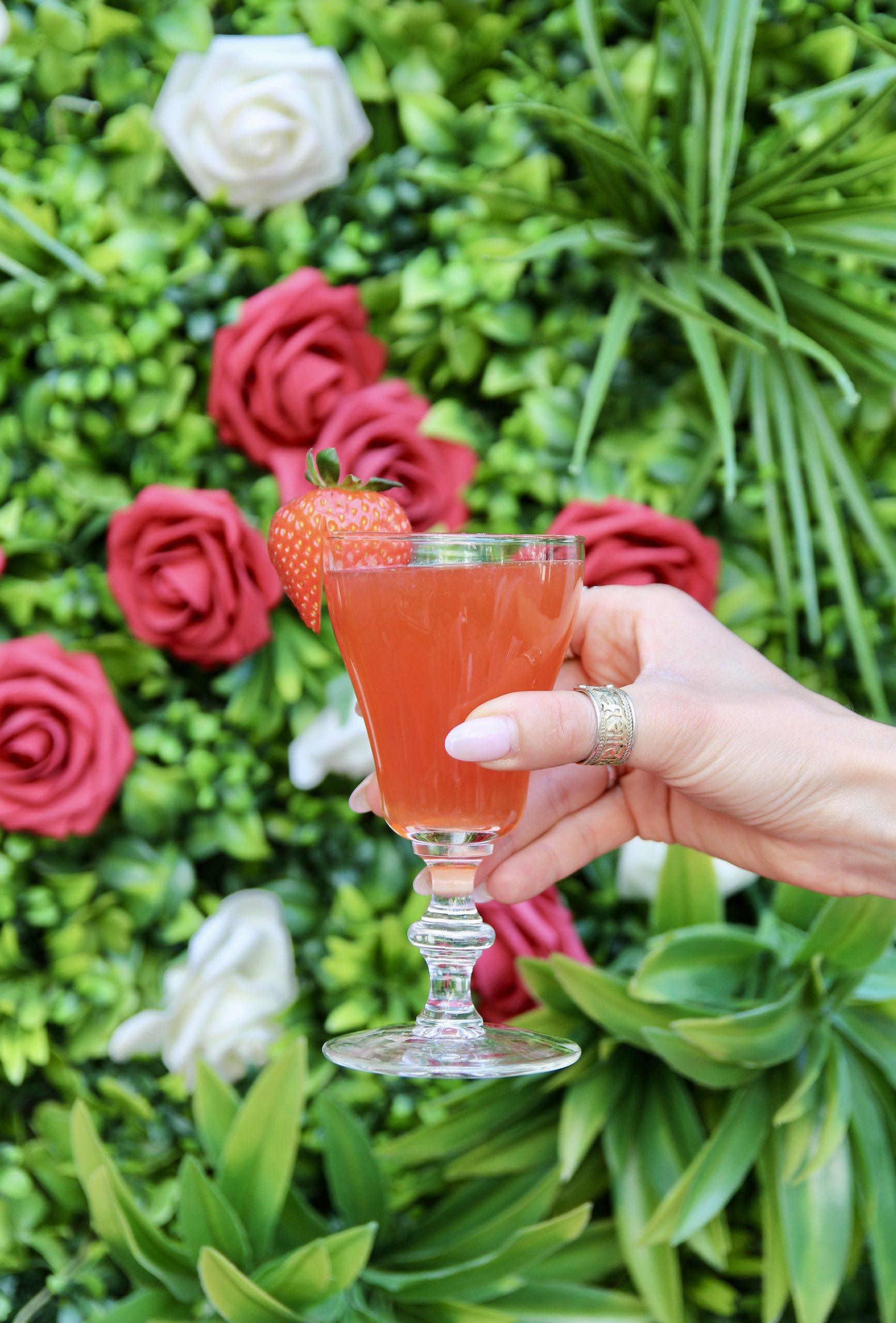A pink cocktail held up against a wall of roses.